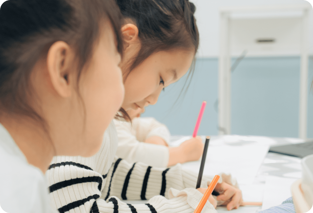 小さな女の子たちが並んで色鉛筆で勉強やお絵かきをしている様子